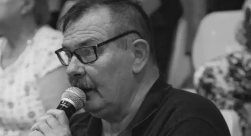Krzysztof Janicki w okularach i z wąsami mówi do mikrofonu, w tle widać ludzi.