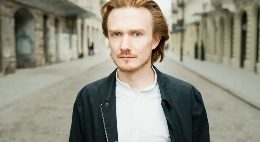 Bartosz Bednarski na tle miejskiej ulicy, z rudymi włosami i zielonymi oczami, w białej koszuli i ciemnej kurtce.
