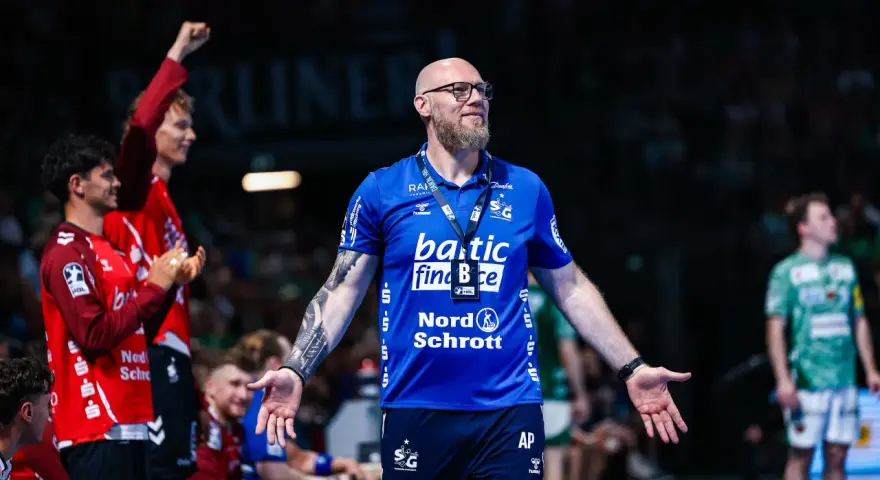 Trener SG Flensburg-Handewitt w niebieskiej koszulce z tatuażem na ramieniu, z otwartymi dłońmi, podczas meczu.