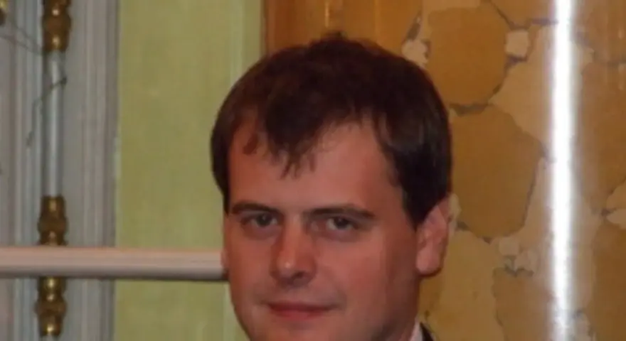 Tomasz Bartoś, zamyślony, w eleganckim stroju, na tle ozdobnej ściany.