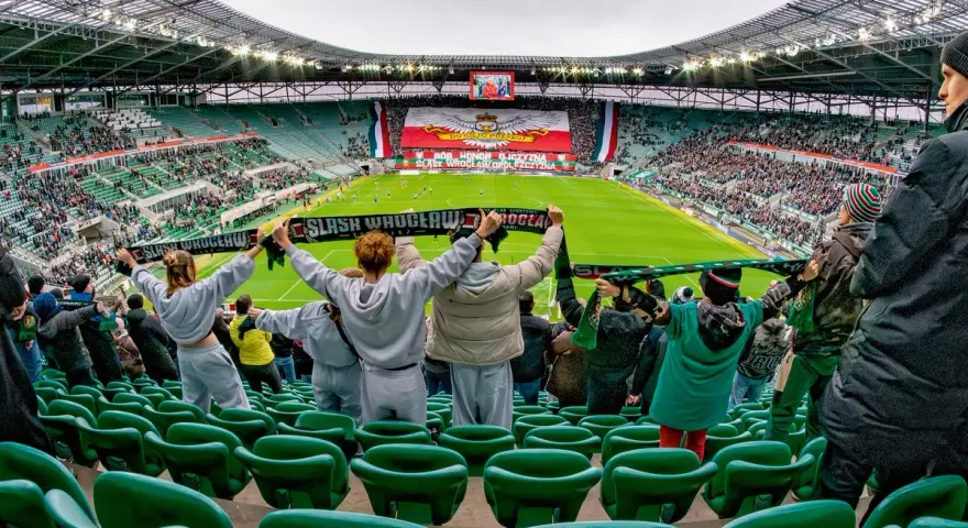 Gwardia Opole kibicuje na stadionie, trzymając szaliki. Na boisku trwa mecz, a trybuny wypełniają fani.