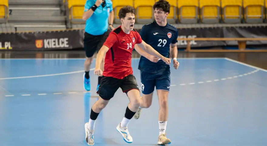 Jakub Smoliński w czerwonej koszulce z numerem 14 biegnie z piłką ręczną, obok zawodnik w granatowej koszulce z numerem 29.