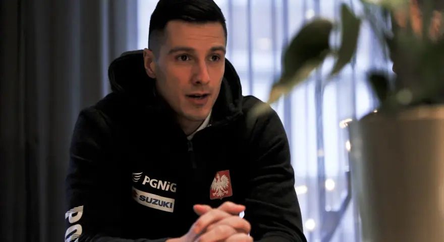 Maciej Pilitowski w czarnej bluzie z orłem i logiem Suzuki, z dłońmi złożonymi na stole, w tle zasłony i rośliny.
