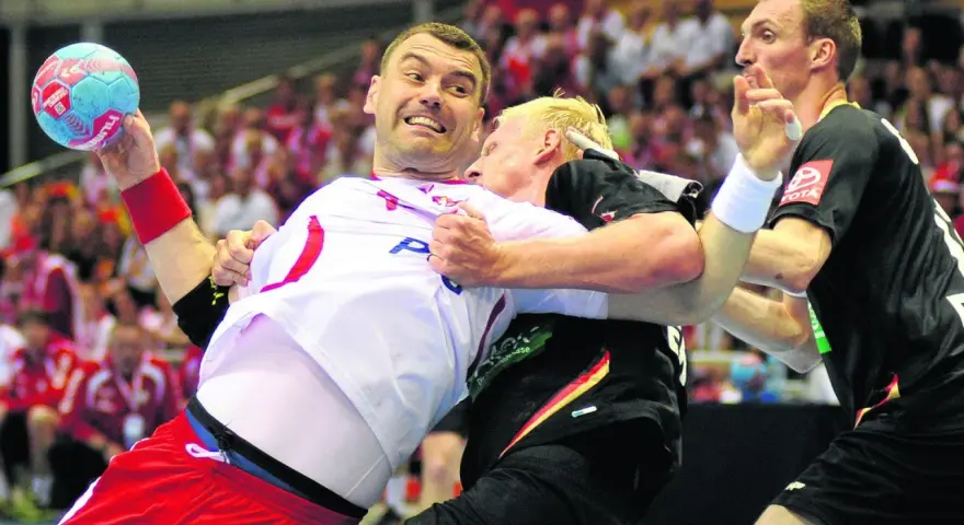 Gracz w białej koszulce z piłką ręczną próbuje wykonać rzut karny, podczas gdy przeciwnik próbuje go zatrzymać.