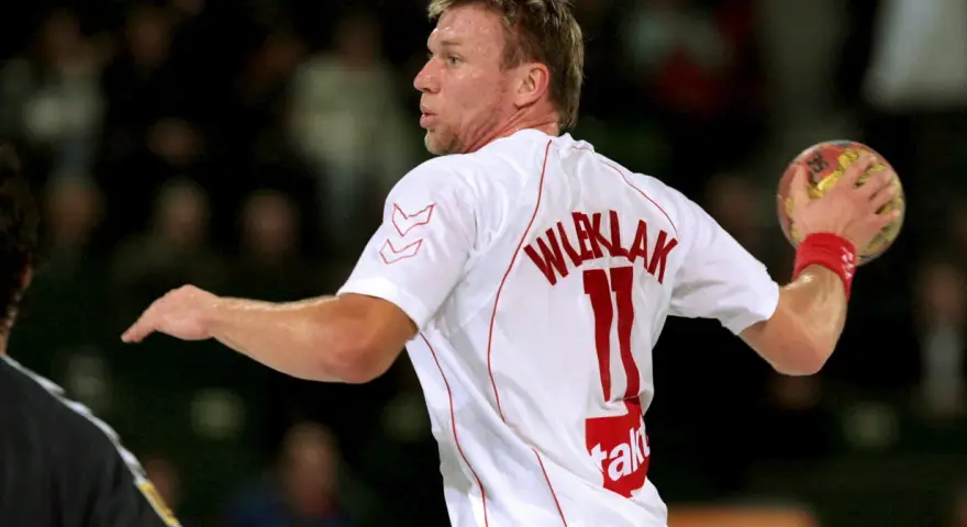 Damian Wleklak, numer 11, w białej koszulce z czerwonymi detalami, przygotowuje się do rzutu piłką ręczną.