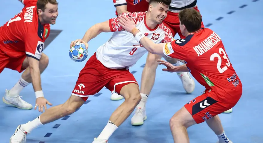 Gracz w białej koszulce z numerem 9 próbuje rzucić piłkę, podczas gdy obrońca w czerwonej koszulce z numerem 23 stara się mu przeszkodzić. Emocjonująca akcja w meczu piłki ręcznej.