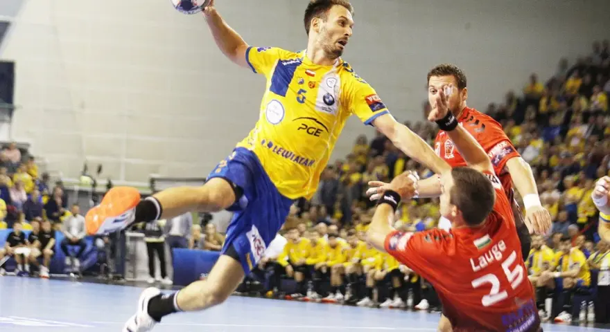 Piłkarz ręczny Telekom Veszprem w żółtej koszulce skacze do rzutu, broniony przez zawodnika w czerwonej.
