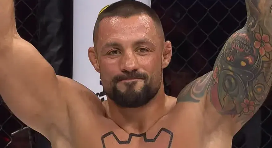 Mateusz Janur, wojownik MMA, z podniesionymi rękami i tatuażami na ramionach i klatce piersiowej, świętuje zwycięstwo.