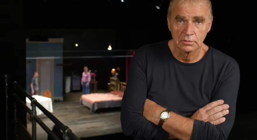 Dawid Głowacki, reżyser teatralny, z rękami skrzyżowanymi na piersi, na tle sceny z łóżkiem i ludźmi.