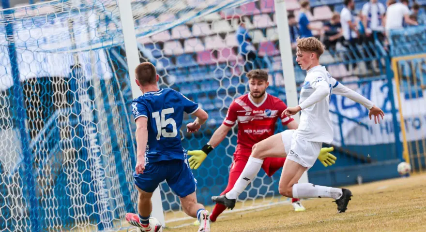 Piłkarz Michał Bartczak w białej koszulce próbuje strzelić gola, obrońca w niebieskiej koszulce z numerem 15 i bramkarz w czerwonej próbuje go zatrzymać.