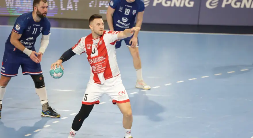 Kamil Adamski w akcji, rzuca piłkę do bramki. Zawodnik w białej koszulce z czerwonymi pasami i numerem 5.