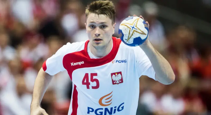 Michał Szyba, numer 45, w białej koszulce z czerwonymi elementami, trzyma piłkę do piłki ręcznej.