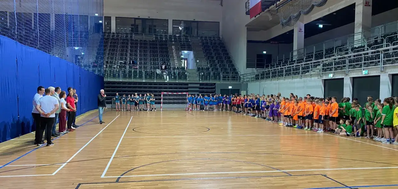 Dwie drużyny dziecięce, ubrane w pomarańczowe i zielone stroje, stoją na hali sportowej przed rozpoczęciem meczu piłka ręczna 1 liga grupa b.