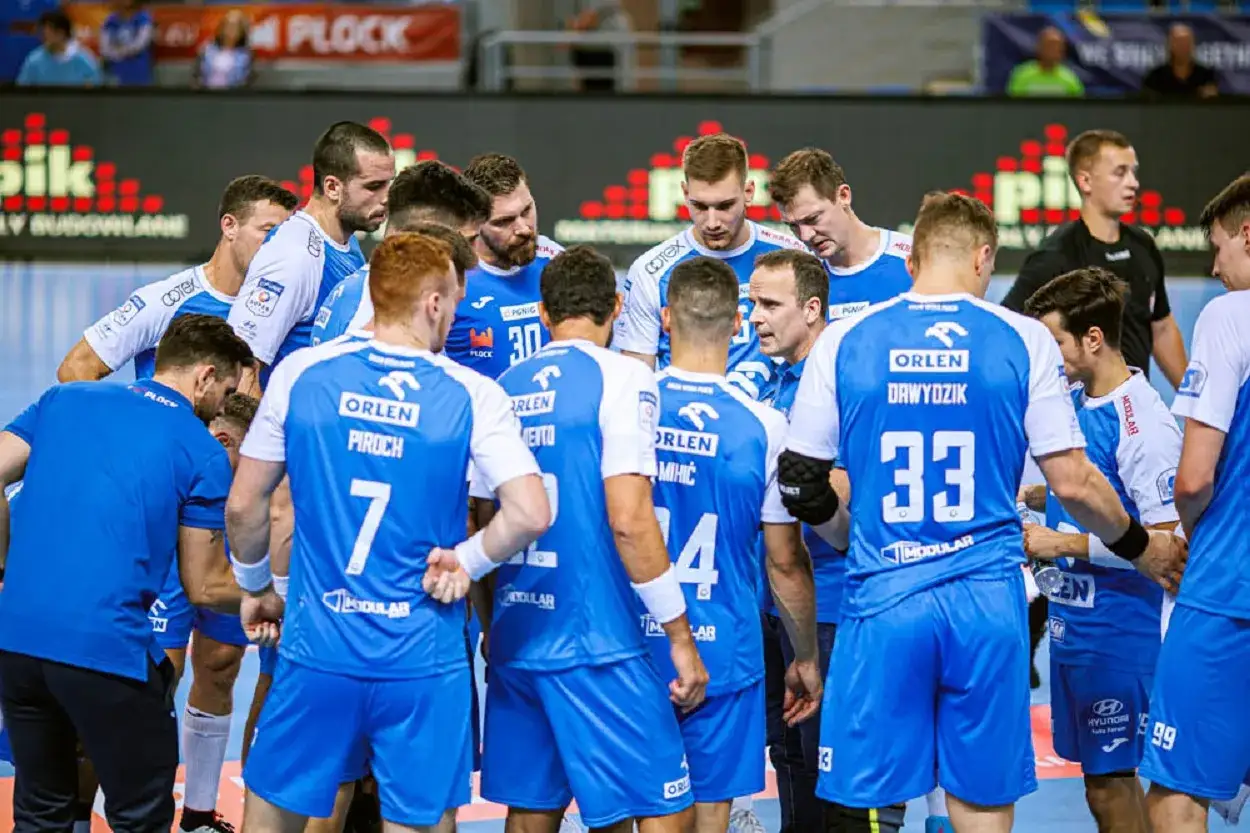 Drużyna gog handball w niebieskich strojach zbiera się na boisku. Zawodnicy z numerami 7, 30, 33 i 4 na plecach.