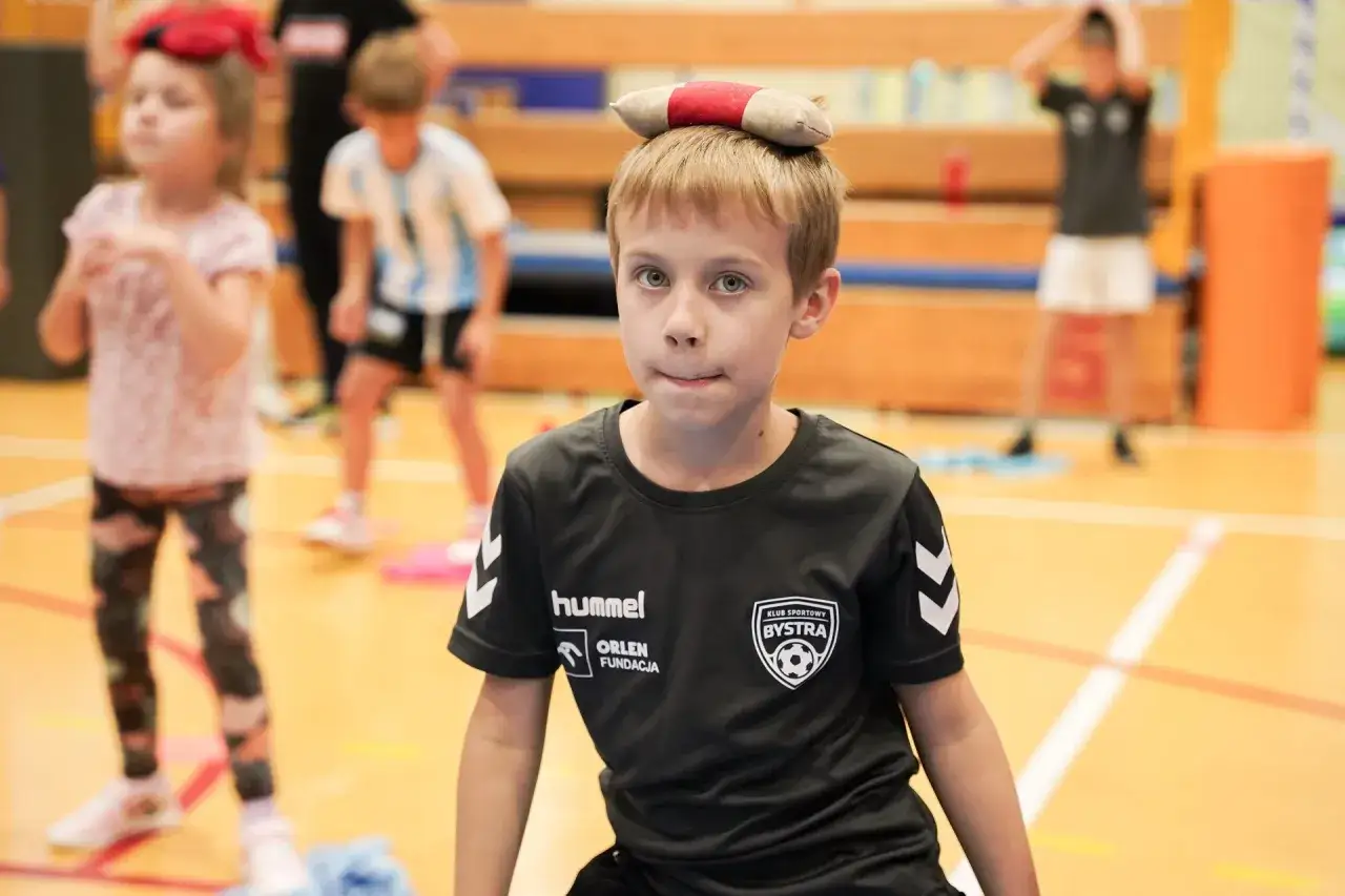 Chłopiec z zabawką na głowie, w koszulce KS Bysstra, podczas zajęć sportowych.