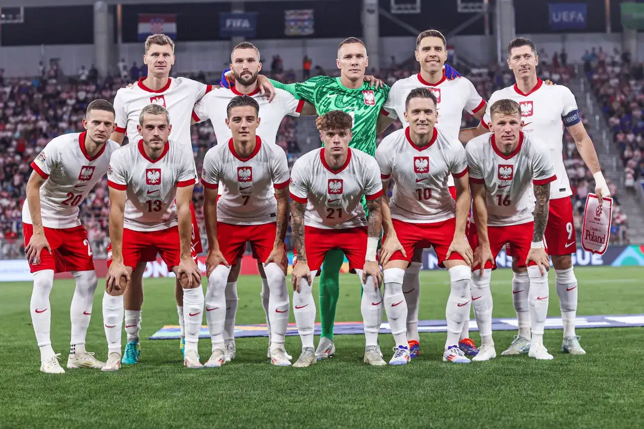 Zgrupowanie kadry Polski na boisku. Piłkarze w białych koszulkach z orłem i czerwonych spodenkach pozują do wspólnego zdjęcia.