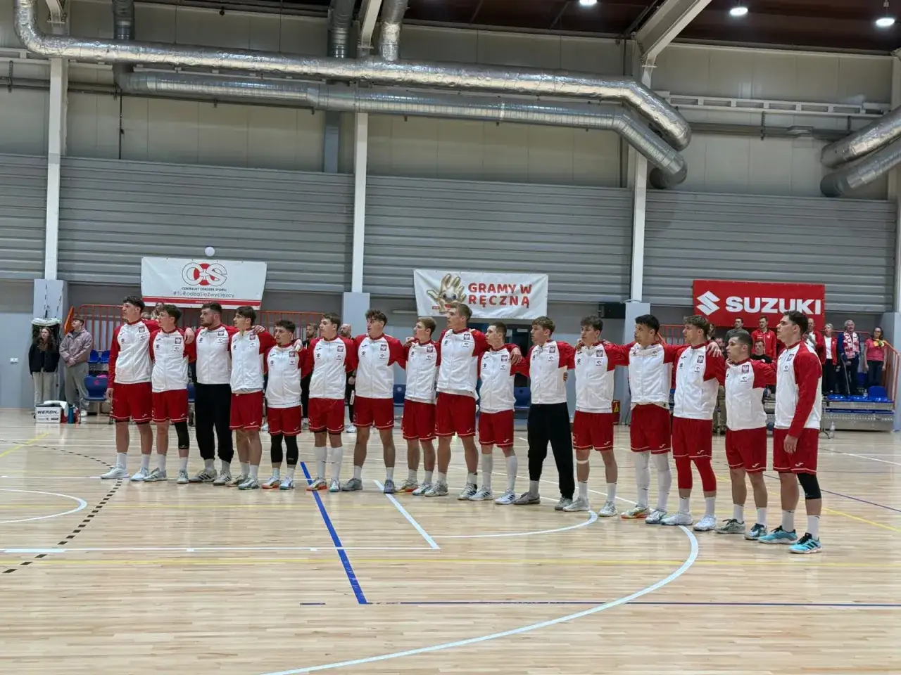 Drużyna młodych piłkarzy, w tym Mateusz Kaczmarczyk, w strojach biało-czerwonych stoi na parkiecie hali sportowej.