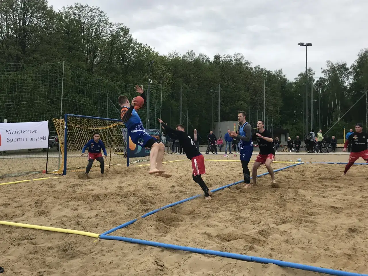 Zawodnicy walczą o piłkę podczas meczu piłki ręcznej plażowej. Jeden z graczy wyskakuje do rzutu, podczas gdy inni próbują go zatrzymać.
