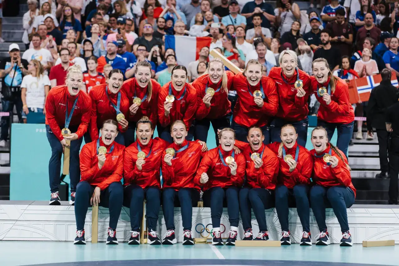 Norweska drużyna piłki ręcznej świętuje zwycięstwo, trzymając złote medale. W tle widać flagę Francji i kibiców.