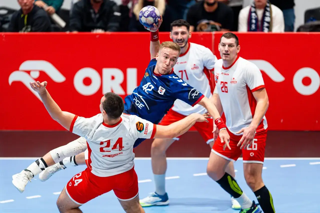 Piłkarz ręczny w skoku rzuca do bramki, broniony przez zawodników w białych strojach z numerami 24 i 26. W tle reklama ORLEN.