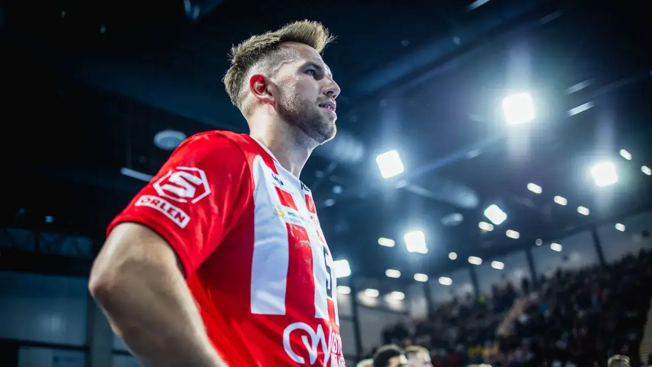 Kamil Adamski w czerwonej koszulce z białymi pasami, na tle hali sportowej z widownią i oświetleniem.