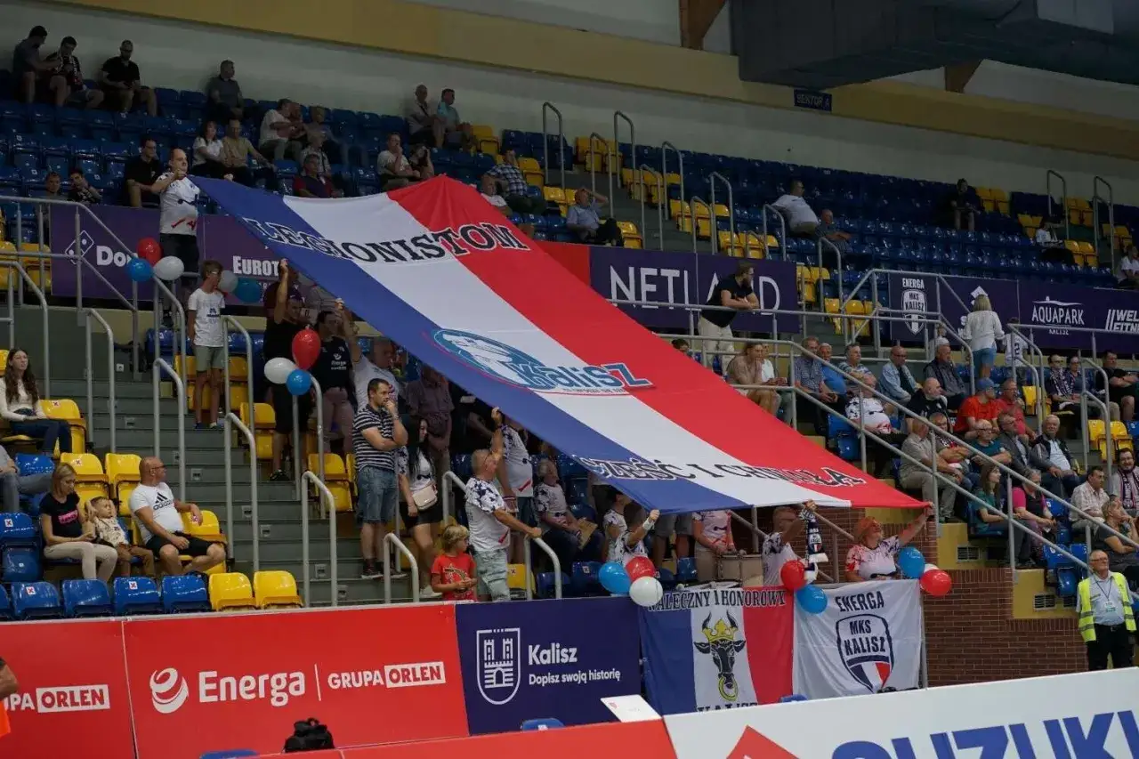 Fani MKS Kalisz rozwijają ogromną flagę na trybunach, kibicując swojej drużynie.