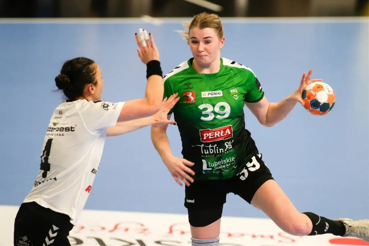 Natalia Nosek w zielonej koszulce z numerem 39 rzuca piłkę do bramki, broni ją przeciwniczka w białej koszulce z numerem 4.