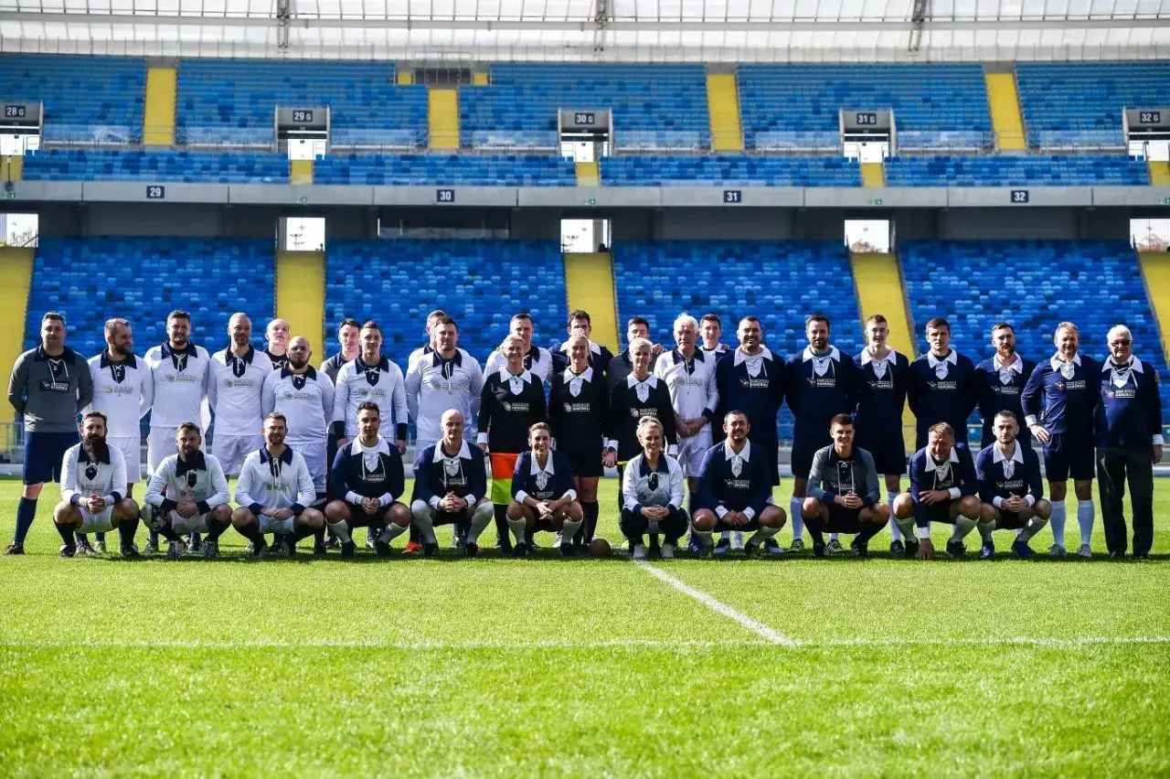 Grupa mężczyzn w strojach sportowych, w tym kilku w białych koszulkach, pozuje na boisku piłkarskim. Czy to drużyna szczypiornista?