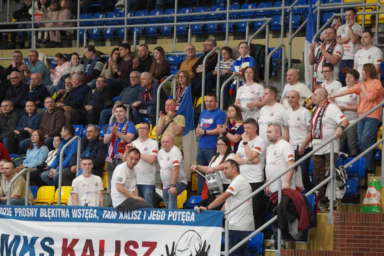 Kibice MKS Kalisz w białych koszulkach i szalikach dopingują drużynę. Na trybunach panuje gorąca atmosfera.
