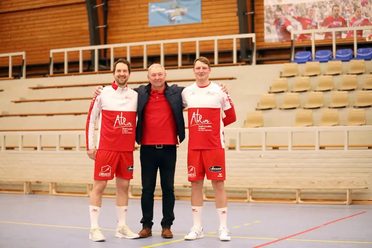 Damian Kędziora z dwoma zawodnikami w strojach sportowych, na tle trybun hali sportowej.