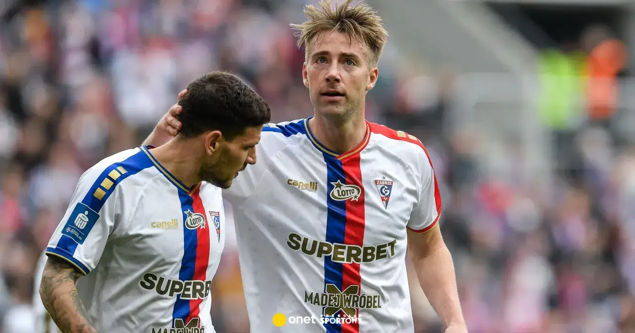 Dw&oacute;ch piłkarzy G&oacute;rnika Zabrze w białych strojach z czerwono-niebieskimi pasami. Jeden z nich, z blond włosami, patrzy przed siebie, drugi, z ciemnymi włosami, ma dłoń na jego ramieniu.