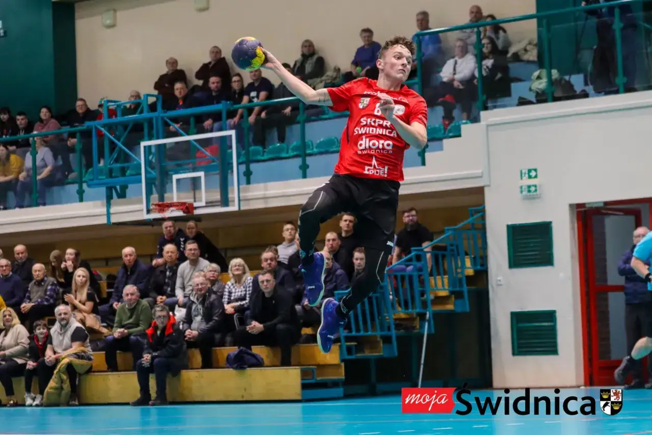 Piłkarz ręczny SKPR Świdnica w skoku do rzutu, z piłką w dłoni, na tle trybun pełnych kibiców.