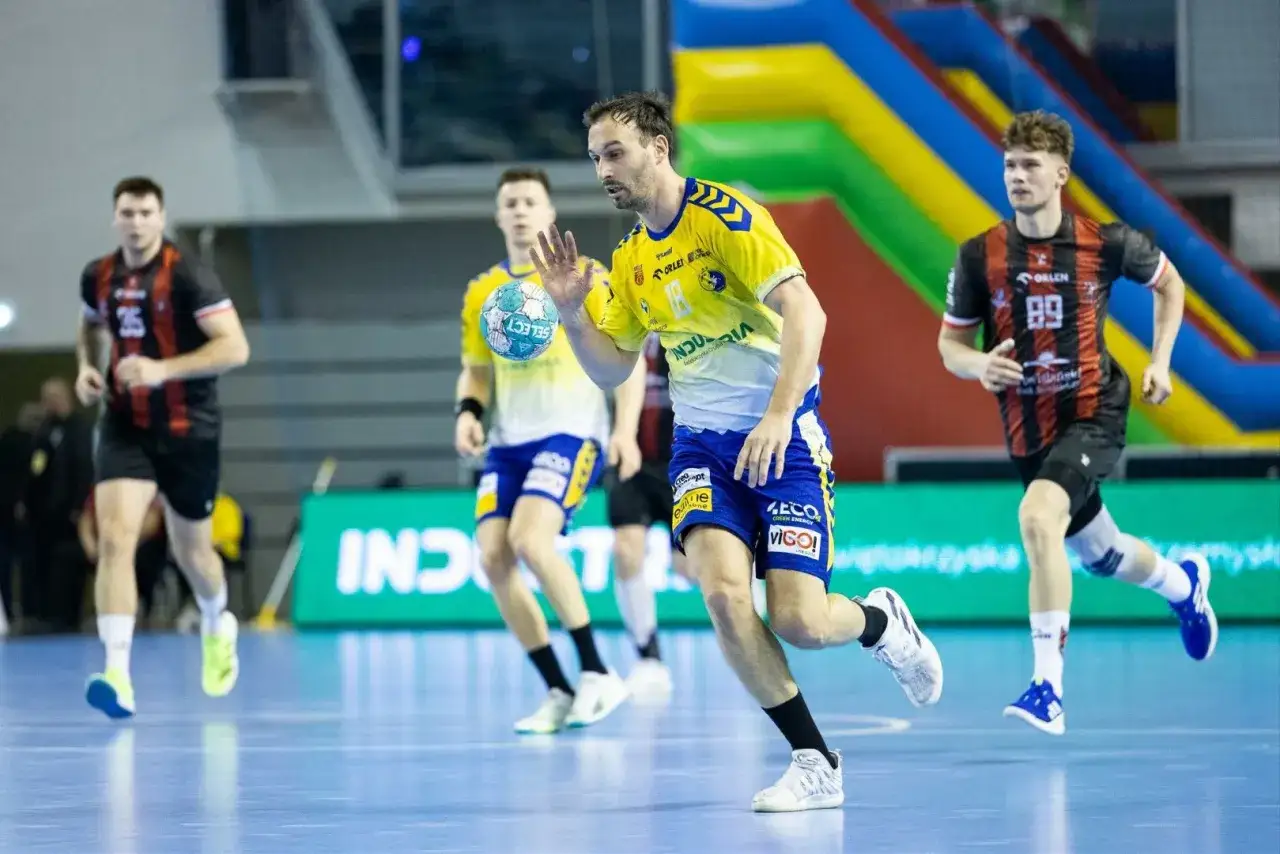 Gracz w ż&oacute;łtej koszulce prowadzi piłkę podczas meczu gog handball. Inni zawodnicy biegną obok.
