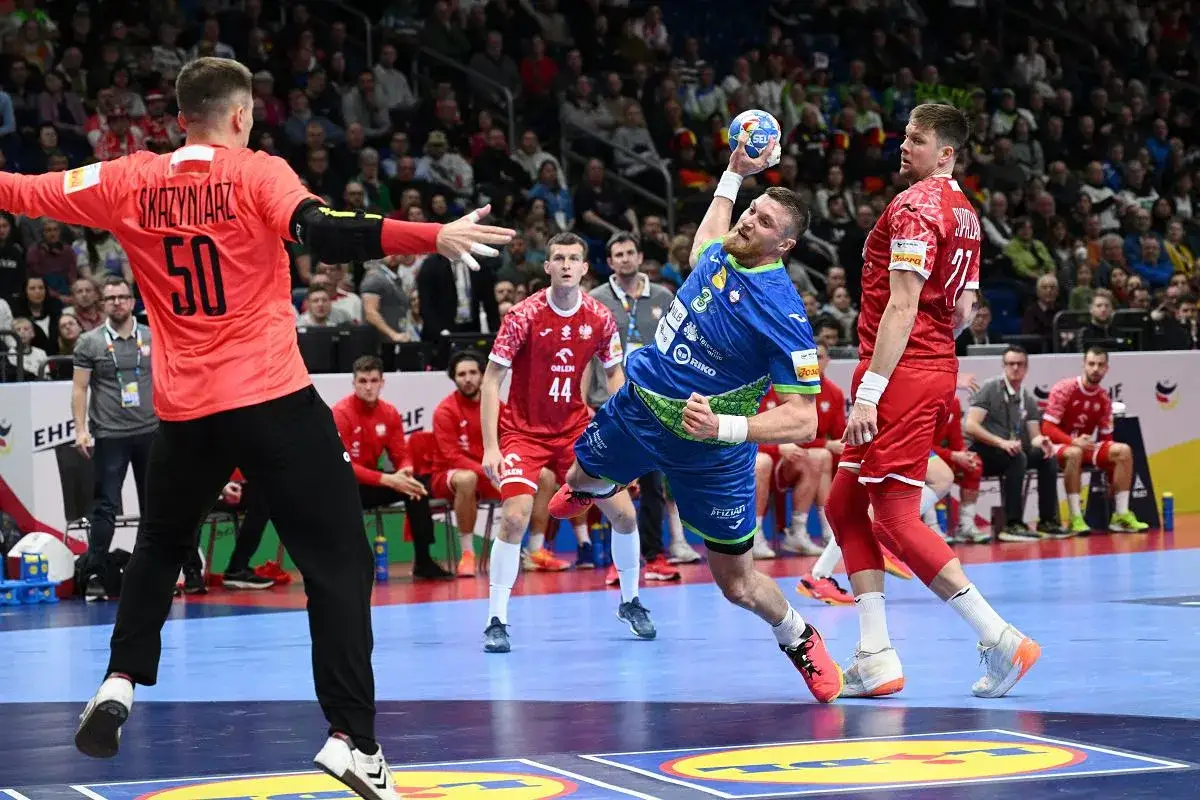 Gracz w niebieskiej koszulce wykonuje rzut w meczu piłki ręcznej. Obok niego bramkarz w czerwonej koszulce z numerem 50. Emocjonująca akcja w piłce ręcznej.