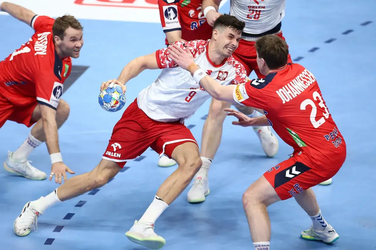 Piłkarz ręczny w białej koszulce z orłem na piersi pr&oacute;buje rzucić piłkę, broniony przez zawodnika w czerwonej koszulce. Emocje w meczu Polska - Słowenia.