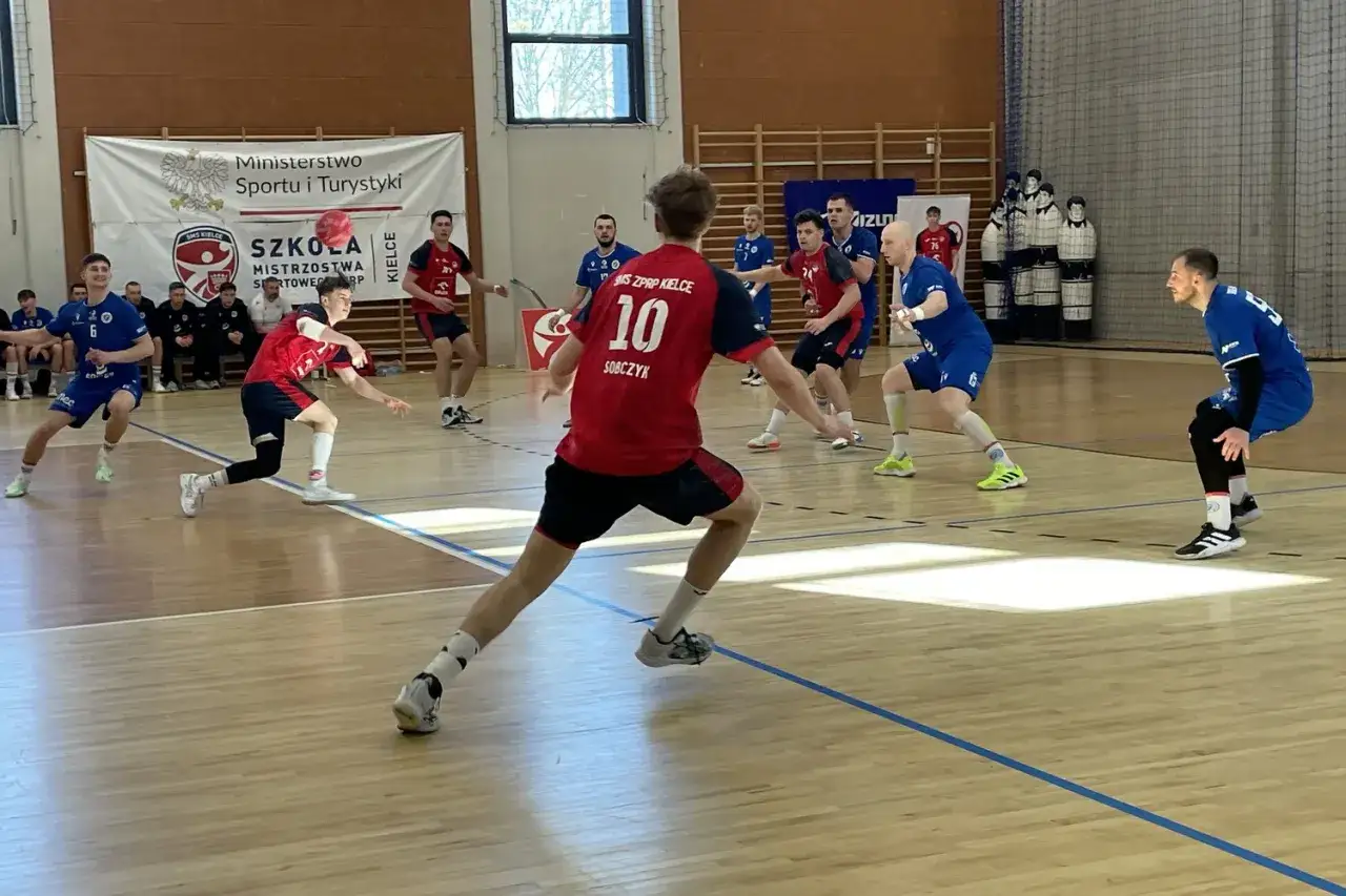 Dynamiczny mecz piłki ręcznej. Zawodnicy w czerwonych i niebieskich strojach walczą o piłkę na parkiecie hali sportowej.