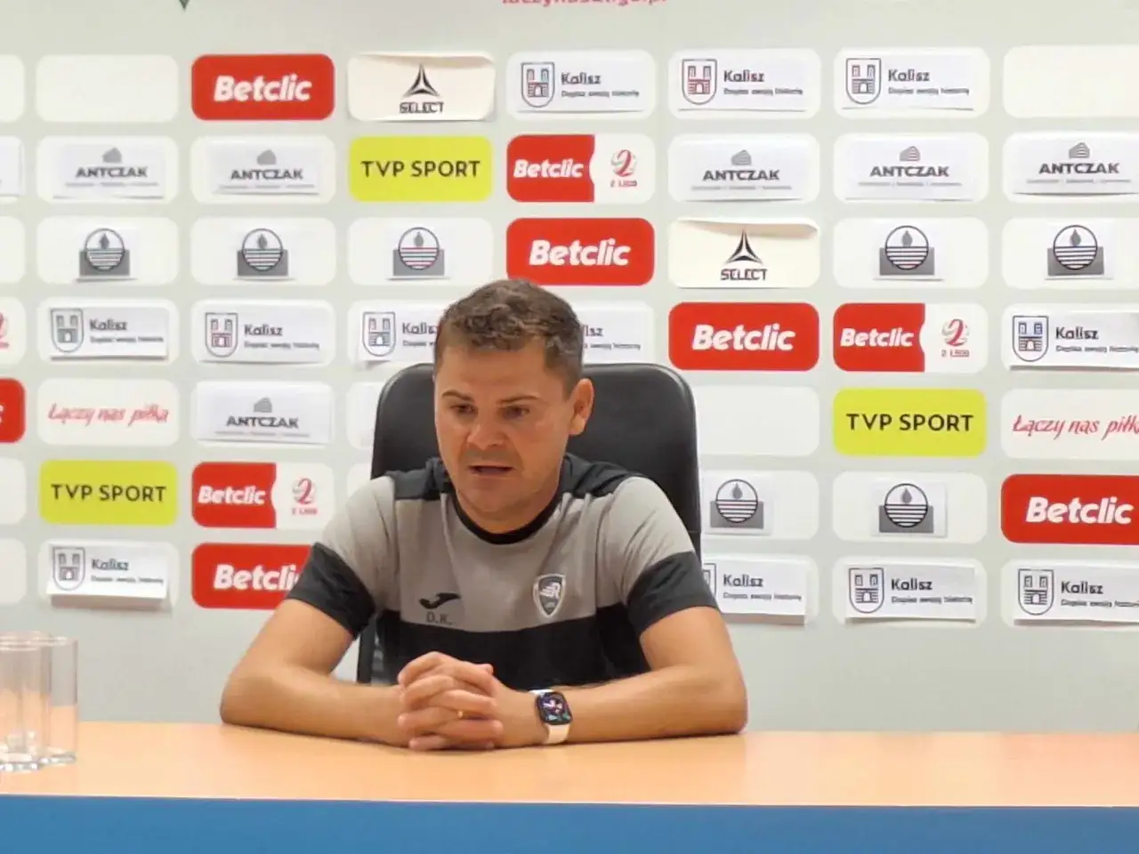 Maciej Kasprzak, trener, siedzi przy stole na tle ścianki z logotypami sponsor&oacute;w, m.in. Betclic i Kalisz.