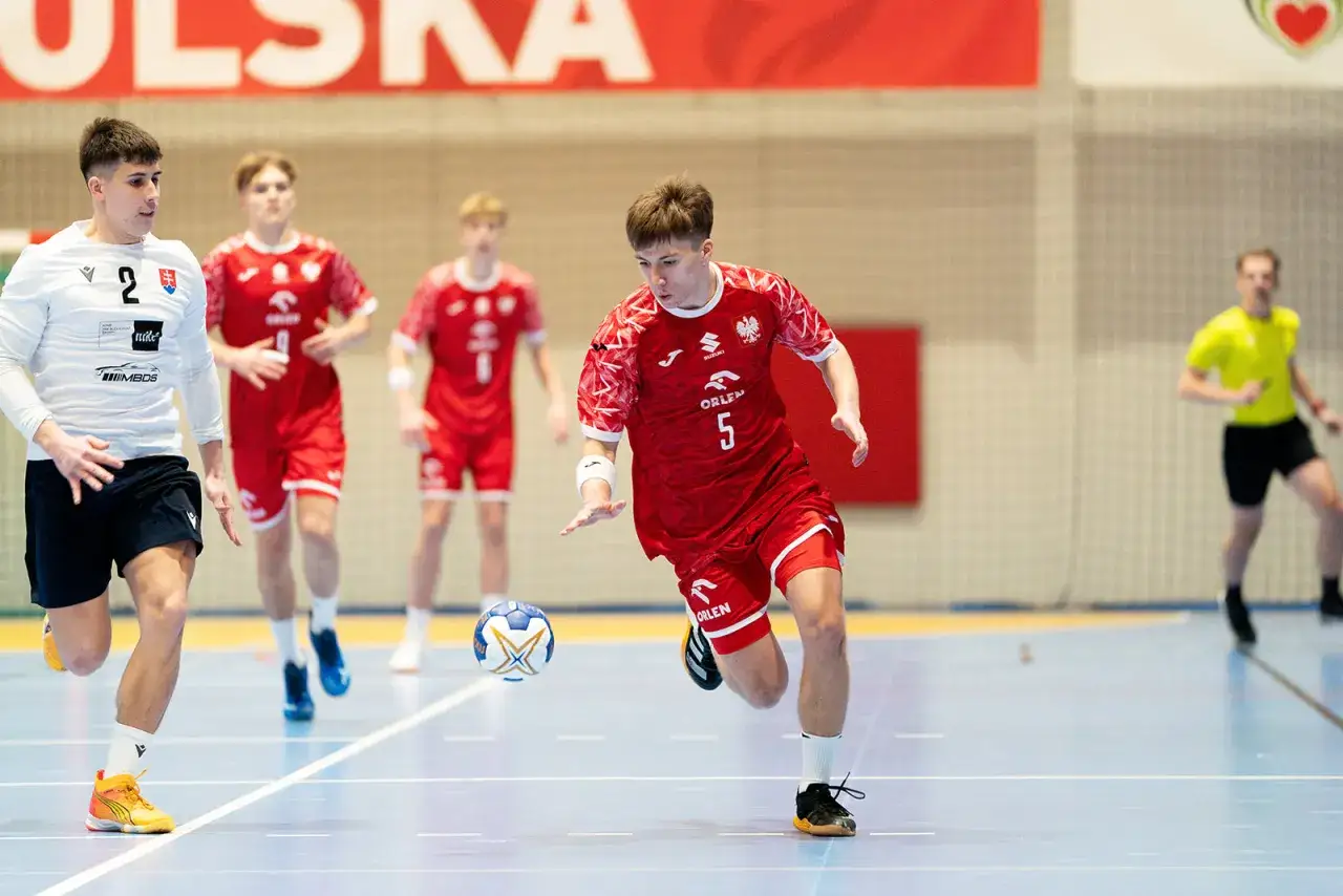 Kamil Skiba w akcji na boisku do piłki ręcznej, w czerwonej koszulce z numerem 5, goni piłkę.