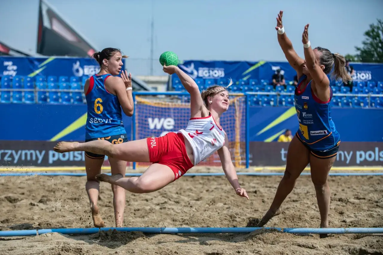 Zawodniczka w skoku po piłkę ręczną plażową, z rywalkami w tle.