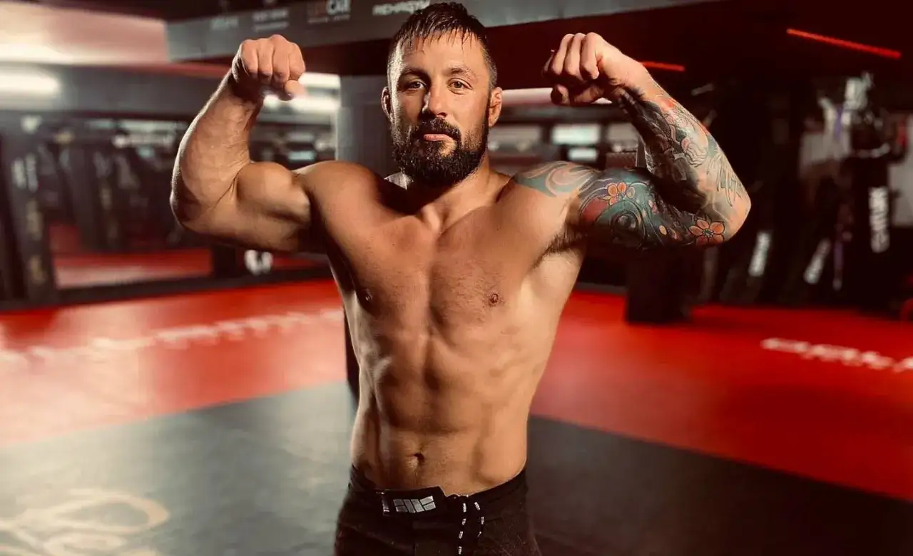Mateusz Janur, muskularny zawodnik MMA, prezentuje swoją formę na macie treningowej, dumnie eksponując tatuaże na ramieniu.