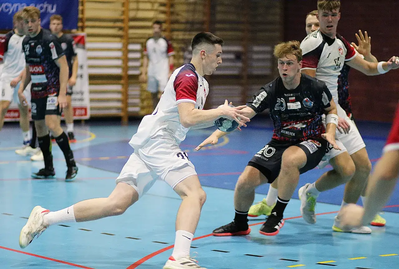 Filip Kwiatkowski w akcji podczas meczu piłki ręcznej. Zawodnik w białej koszulce z czerwonymi rękawami próbuje rzucić piłkę, podczas gdy przeciwnik w czarnej koszulce go blokuje.