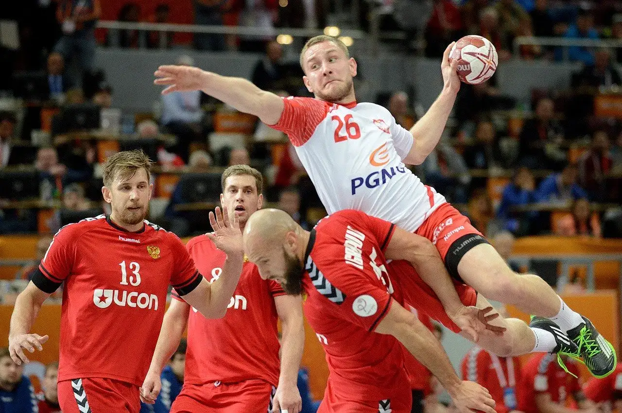 Gracz w białej koszulce z numerem 26, reprezentujący Polskę w piłce ręcznej, wyskakuje do rzutu, podczas gdy obrońcy w czerwonych strojach pr&oacute;bują go zatrzymać.
