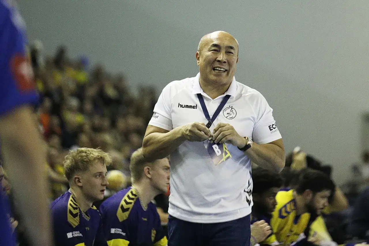 Uśmiechnięty trener Vive Kielce w białej koszulce z logo Hummel, z zawodnikami w tle.