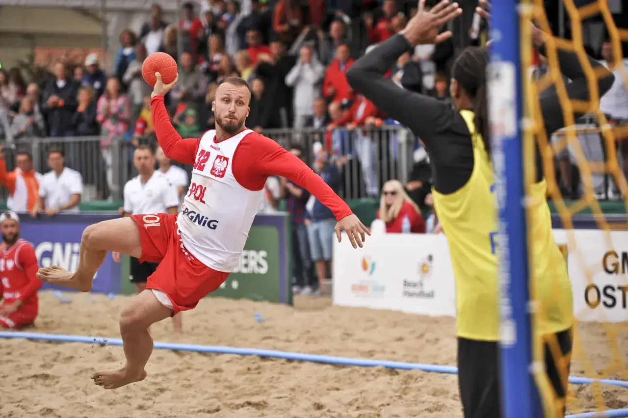 Mateusz Orłowski w skoku rzuca piłką do bramki podczas meczu piłki ręcznej plażowej.