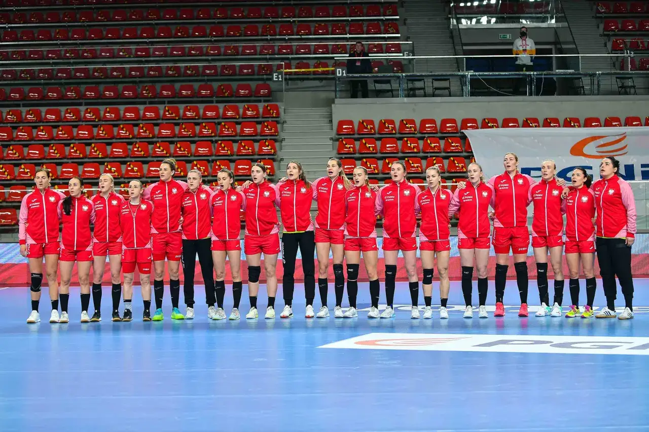 Drużyna piłki ręcznej kobiet w czerwonych strojach z orzełkiem na piersiach, stojąca przed trybunami.
