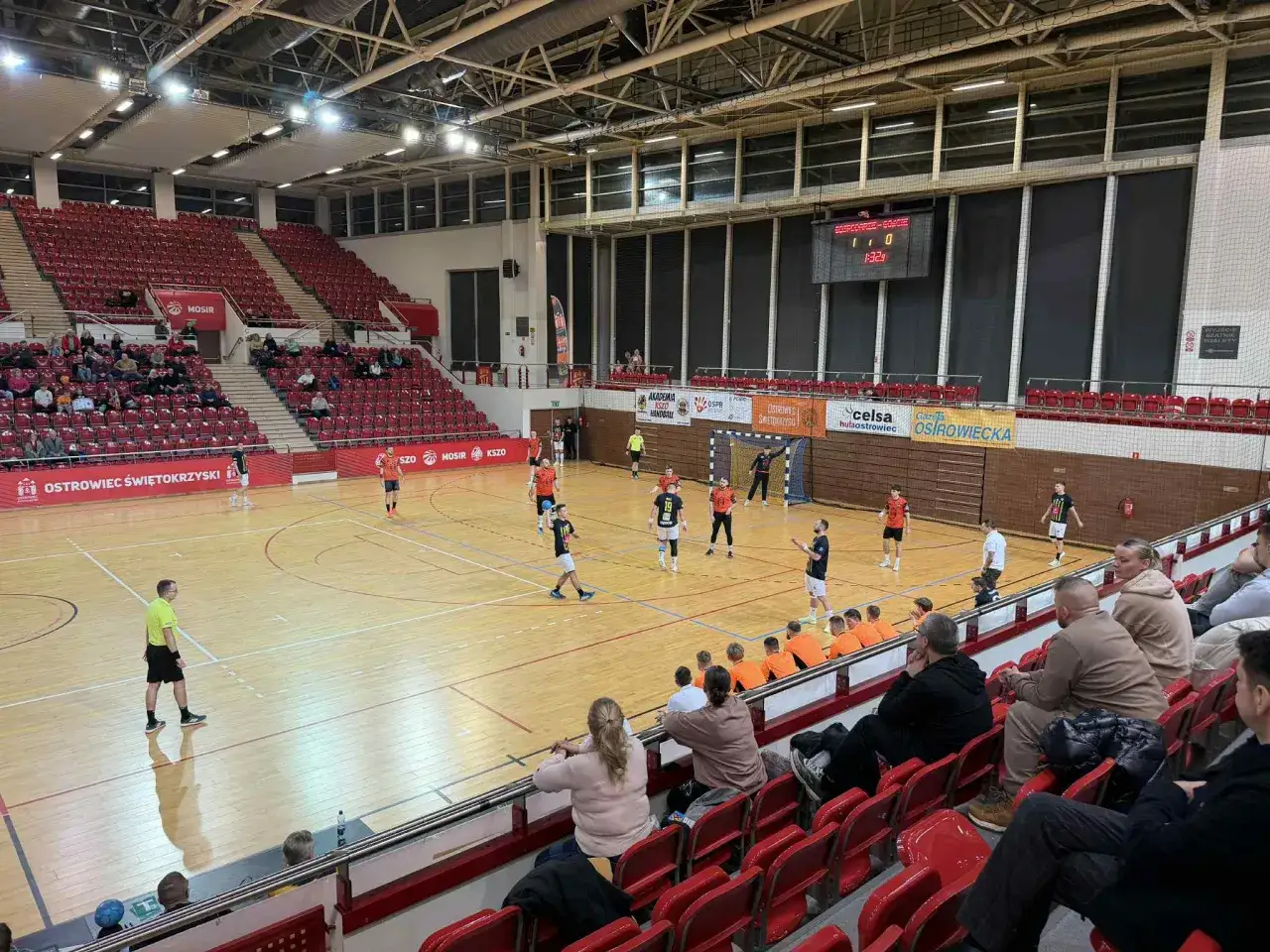 Mecz kszo handball w hali sportowej. Zawodnicy na boisku, kibice na trybunach. Wynik 1:0.