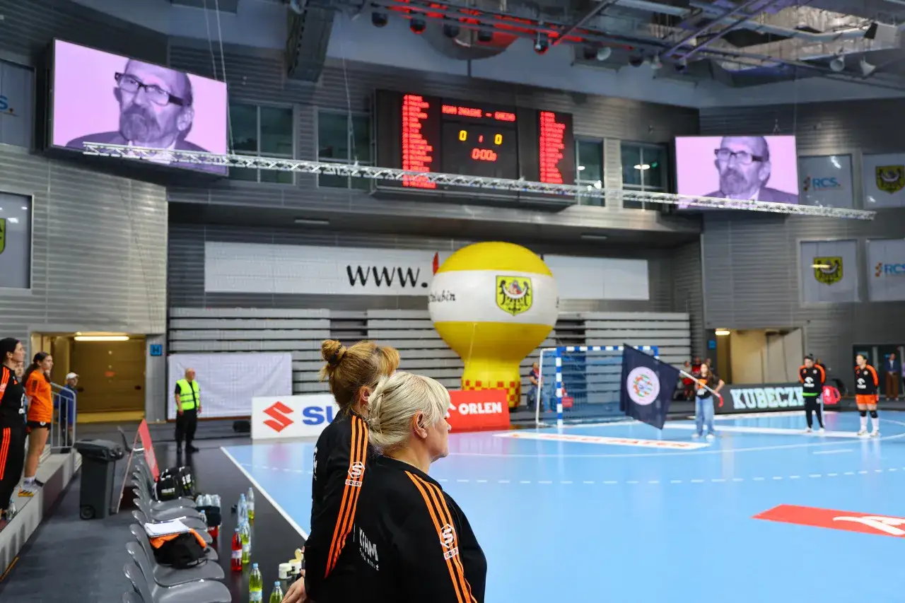 Kobiety w strojach MKS Urbis Gniezno patrzą na boisko. Na ekranach widać mężczyzn, a na tablicy wyników 0:0.