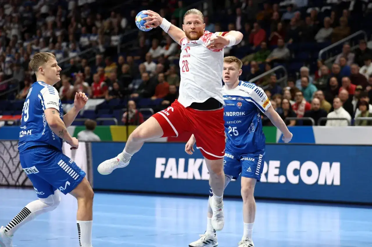 Piłkarz ręczny w skoku rzuca piłkę, obrońcy próbują go zatrzymać. Dynamiczna akcja w meczu piłki ręcznej mężczyzn.