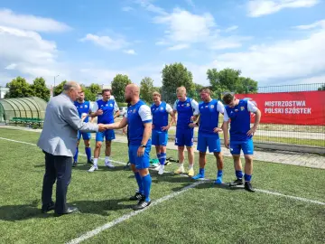 Starszy pan wita drużynę Grunwald Ruda Śląska na boisku. Piłkarze w niebieskich strojach czekają na rozpoczęcie meczu.