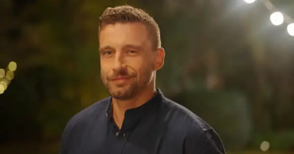 Uśmiechnięty Jakub Wojnowski w granatowej koszuli, w tle rozmyte światła i zieleń.